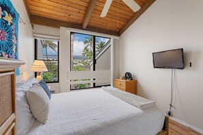 Ein Schlafzimmer mit einem Bett, einem an der Wand montierten Fernseher, einem Fenster mit Blick auf Palmen und einer Holzbalkendecke.