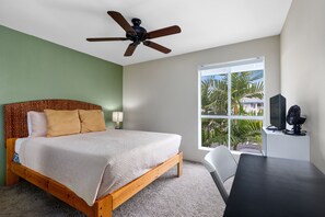 Un dormitorio con cama de madera, ventilador de techo, escritorio con silla y ventana con vista a palmeras.