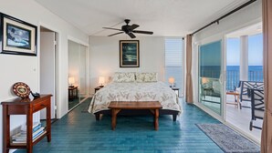 A bedroom with a bed, a ceiling fan, a nightstand, a bedside table, and a view of the ocean through a sliding glass door. - Alii Villas #232 2 Bedroom Condo (Kailua-Kona)