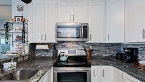 A modern kitchen with white cabinets, a stainless steel microwave, a stove, and an oven. There is a granite countertop, a sink, and a backsplash with a mosaic tile design. - Alii Villas #232 2 Bedroom Condo (Kailua-Kona)