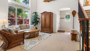 A spacious living room with a large window, a wooden cabinet, a wicker chair, and a staircase. - Fairways Mauna Lani 1406 2 Bedroom Condo (Kamuela)