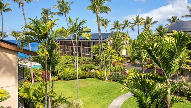 A hotel with a lush garden, palm trees, and a view of the ocean. - Kona Makai 3301 2 Bedroom Condo (Kailua-Kona)