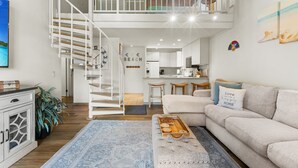 A modern living room with a staircase, a sofa, a coffee table, and a kitchen area in the background. - Kona Makai 3301 2 Bedroom Condo (Kailua-Kona)