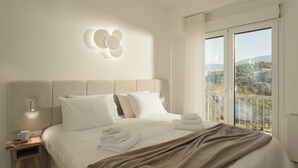 Un dormitorio moderno con una cama grande, una mesita de noche, una lámpara de pared y vistas a un paisaje urbano desde un balcón.