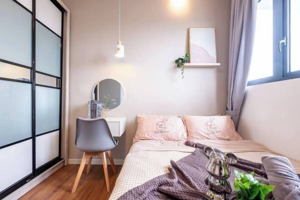 A bedroom with a bed, a desk, a chair, a hanging light, a wall-mounted shelf with a plant, and a window with curtains.