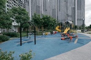 A playground with swings, slides, and climbing structures surrounded by greenery and tall buildings.