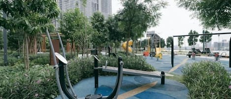 An outdoor exercise area with a stationary bike and elliptical machine, surrounded by greenery and a paved path.