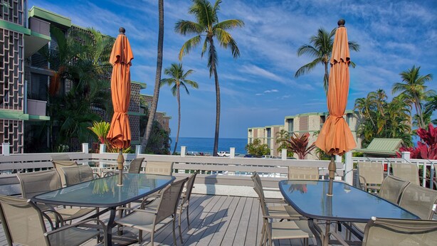 An outdoor dining area with tables and chairs, surrounded by palm trees and a view of the ocean. - Alii Villas #301 1 Bedroom Condo (Kailua-Kona)