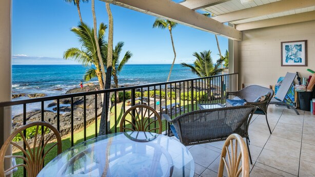 A balcony with a glass table and wicker chairs, overlooking a beach with palm trees and the ocean. - Kona Bali Kai #219 2 Bedroom Condo (Kailua-Kona)