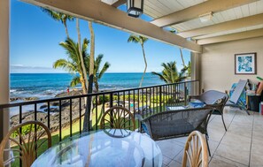 Ein Balkon mit Glastisch und Korbstühlen, der Blick auf einen Strand mit Palmen und dem Meer freigibt.