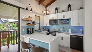 A modern kitchen with white cabinets, a central island, and a view of a balcony with plants. - Kona Makai 3302 2 Bedroom Condo (Kailua-Kona)