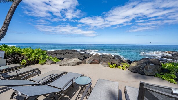 A beachside lounge with lounge chairs and a table, overlooking the ocean with rocky terrain and a clear sky. - Kona Makai 3302 2 Bedroom Condo (Kailua-Kona)