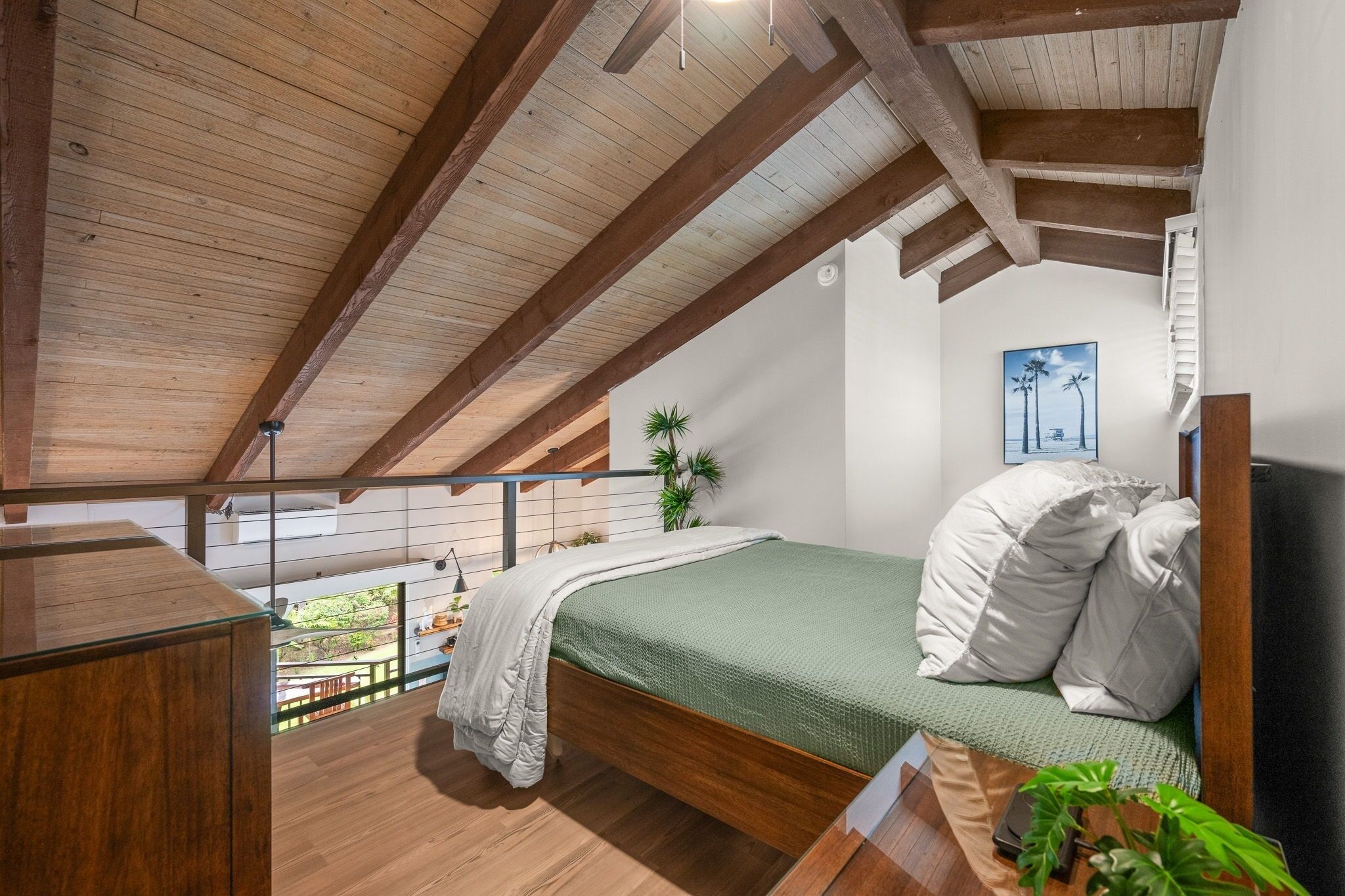 A bedroom with a wooden ceiling, a bed with white bedding, a small plant, and a painting on the wall.
