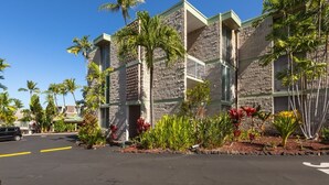 A building with a stone facade and multiple balconies, surrounded by tropical plants and palm trees.