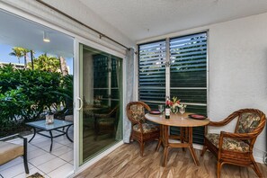 A patio area with a round table, two wicker chairs, and a glass door leading to a balcony. - Alii Villas #109 1 Bedroom Condo (Kailua-Kona)