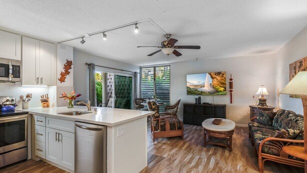 A modern kitchen with white cabinets, a dishwasher, and a microwave. There is a dining area with a wooden table and chairs. The living room features a couch and a painting on the wall.