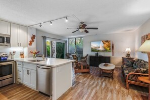 A modern kitchen with white cabinets, a dishwasher, and a microwave. There is a dining area with a wooden table and chairs. The living room features a couch and a painting on the wall. - Alii Villas #109 1 Bedroom Condo (Kailua-Kona)