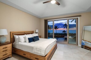 A bedroom with a large bed, a nightstand, a lamp, and a view of the beach through a sliding glass door.