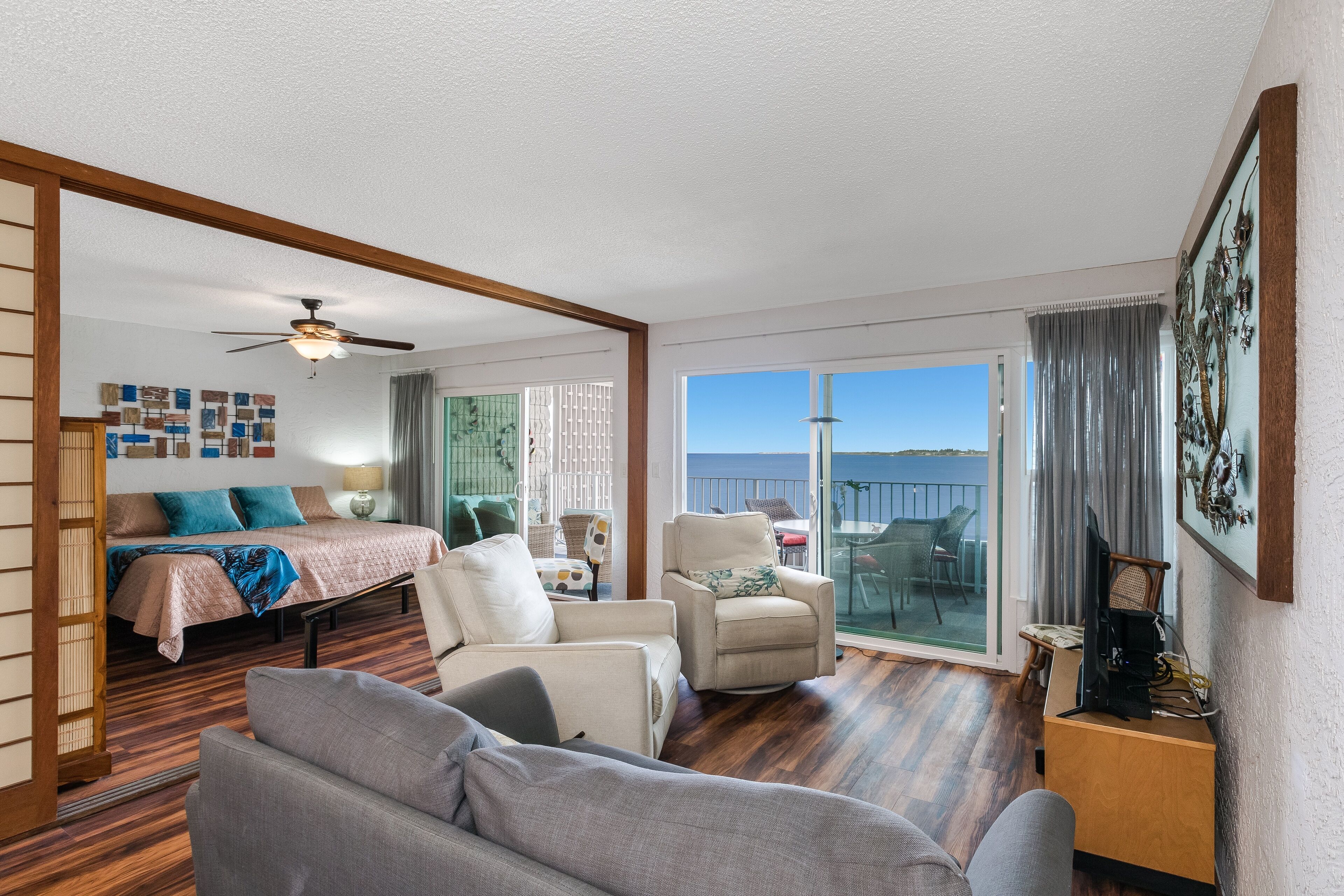 A spacious living area with a large bed, a sofa, a chair, and a TV stand. There is a balcony with a view of the sky.