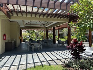 A covered outdoor area with tables and chairs, a fire extinguisher, and a view of a garden and buildings in the background. - Waikoloa Beach Villas #o23 2 Bedroom Condo (Waikoloa)