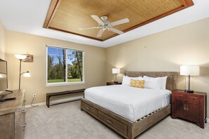 A bedroom with a large bed, wooden headboard, bedside lamps, a ceiling fan, a window with a view of trees, and a desk with a lamp. - Waikoloa Beach Villas #o23 2 Bedroom Condo (Waikoloa)