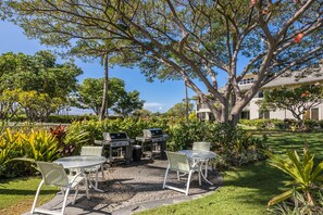 An outdoor patio with a grill, chairs, and a table surrounded by greenery and a large tree. - Waikoloa Beach Villas #o23 2 Bedroom Condo (Waikoloa)