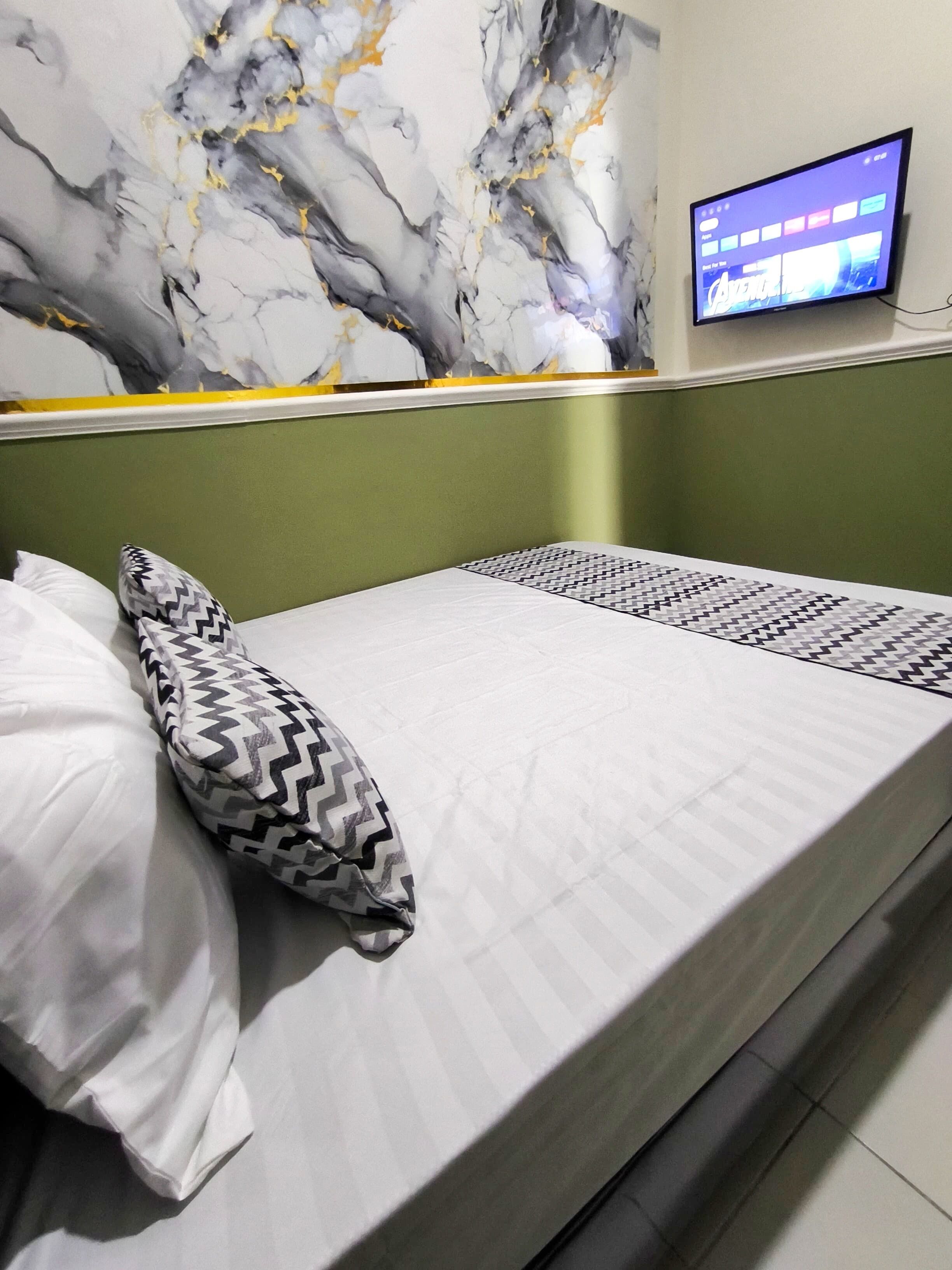 Una camera d'albergo con un letto, un cuscino decorato, una televisione a parete e un rivestimento murale simile al marmo.