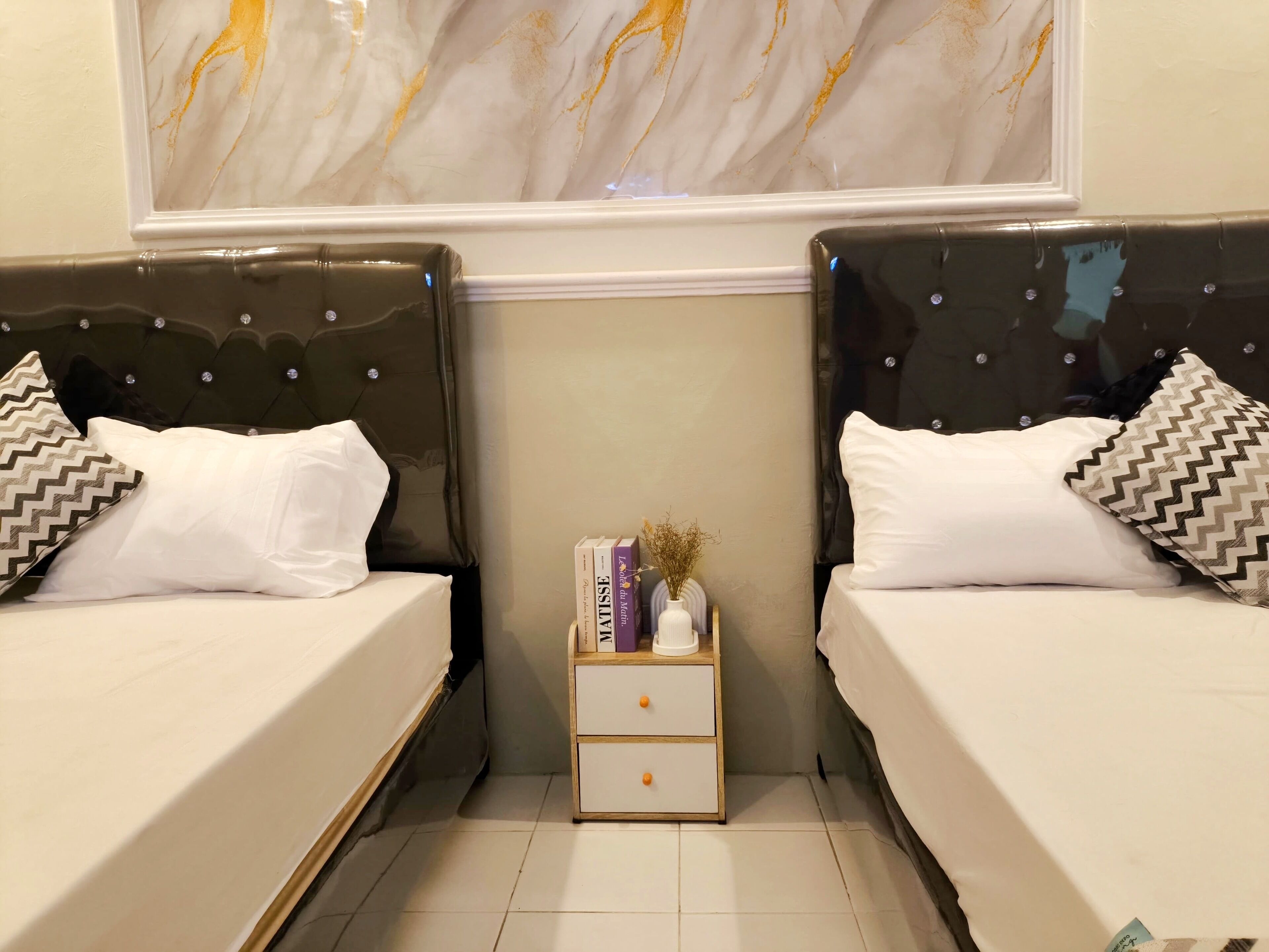 A double room with two beds, a nightstand with books and a vase, and a marble wall above.