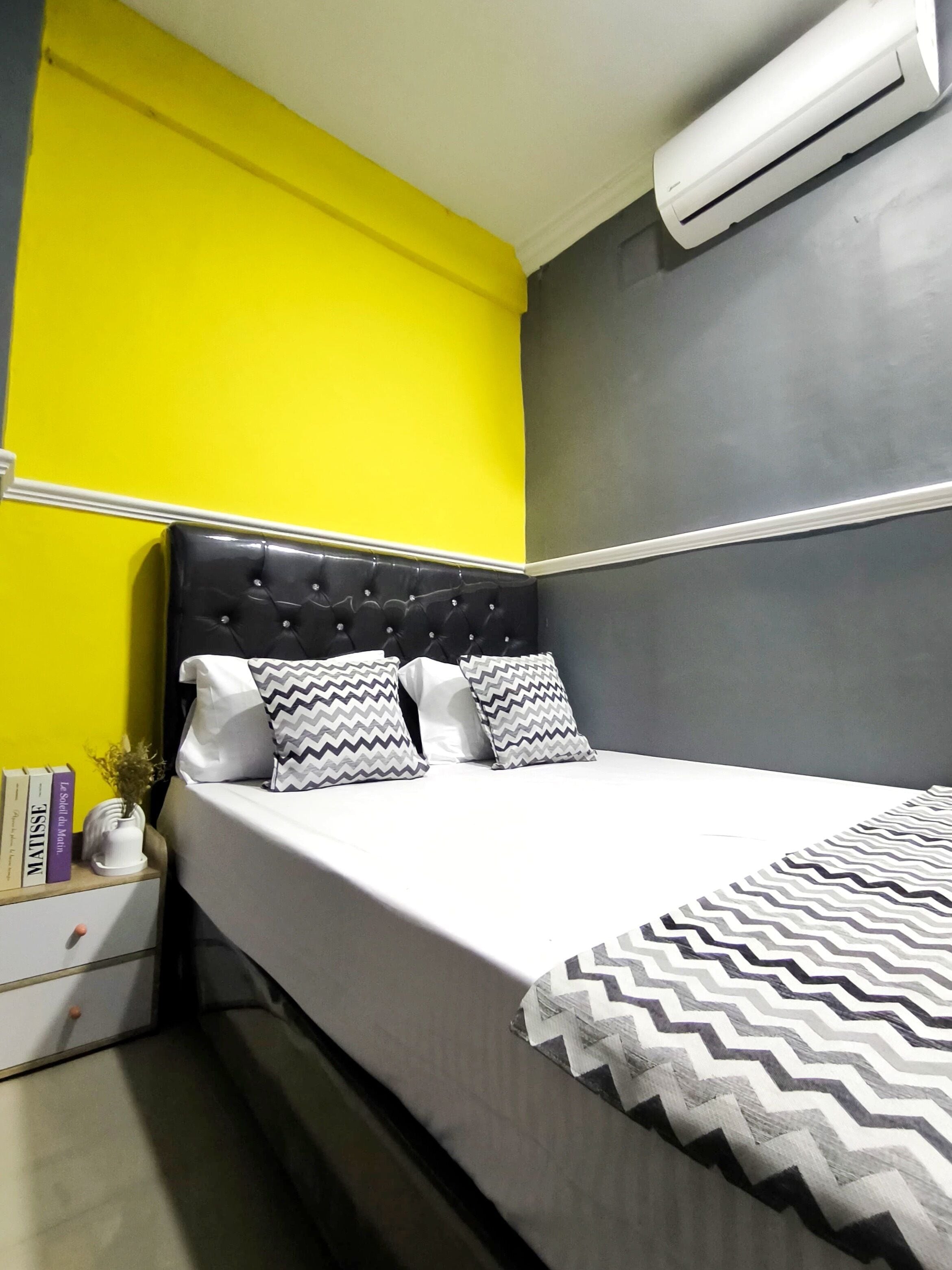 A bed with a white mattress and grey headboard, two pillows with zigzag patterns, a grey wall, and an air conditioning unit above the bed.