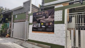 A residential building with a banner advertising a property named "Alap To Stay." The banner lists amenities such as an AC, outdoor bathroom, shared kitchen, Android TV, WiFi internet, and a smoking area.