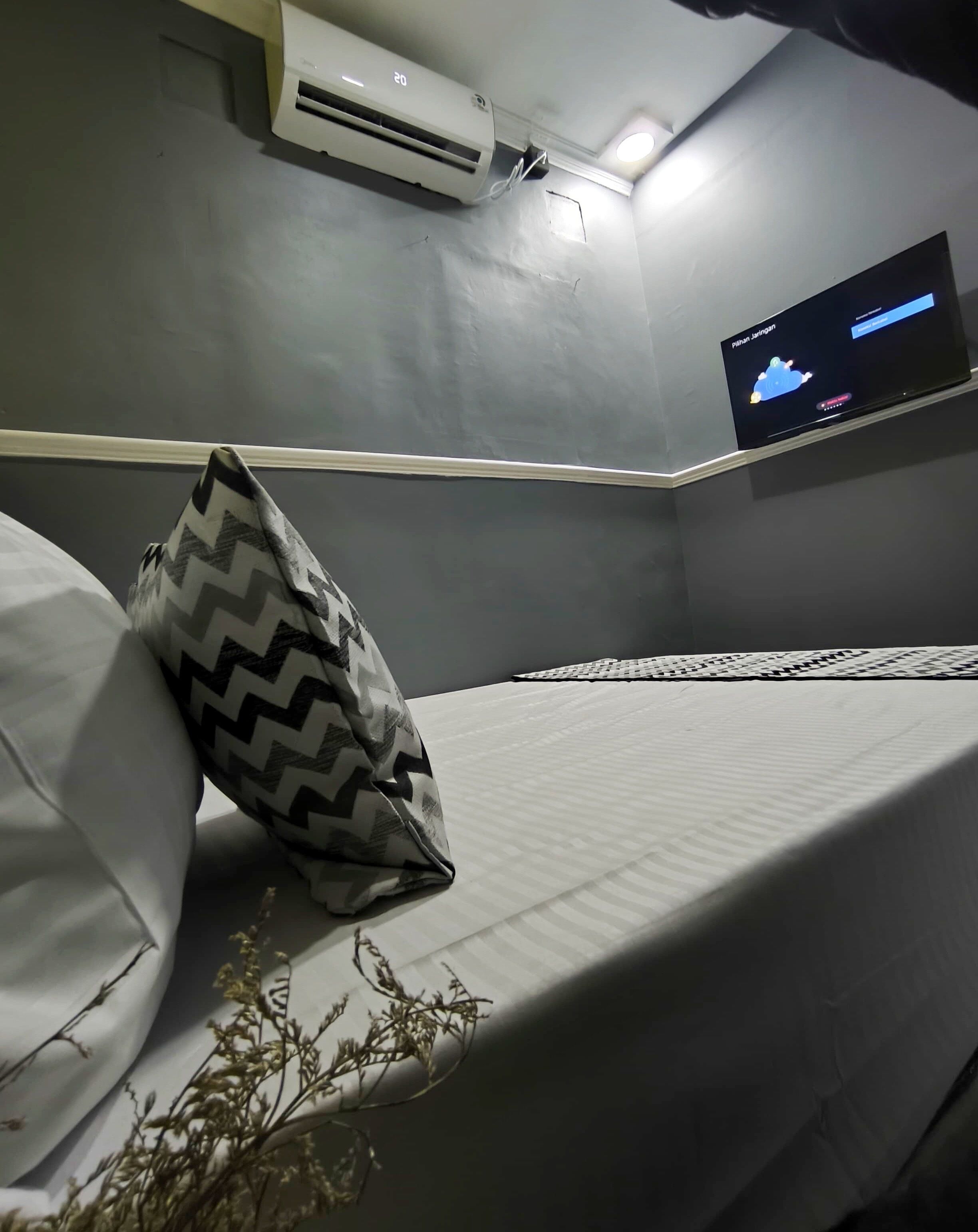 Una habitación con una cama, un cojín estampado, un televisor montado en la pared y un equipo de aire acondicionado.