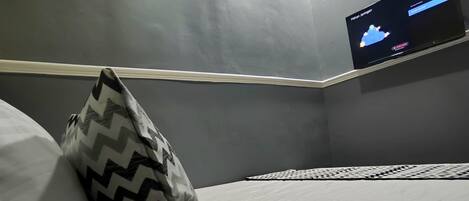Una habitación con una cama, un cojín estampado, un televisor montado en la pared y un equipo de aire acondicionado.