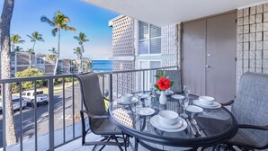 A balcony with a dining table set for four, overlooking a parking area and palm trees, with a view of the ocean and buildings in the background. - Alii Villas #224 1 Bedroom Condo (Kailua-Kona)