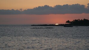 A sunset over a calm sea with a rowboat and a distant shoreline with palm trees. - Alii Villas #224 1 Bedroom Condo (Kailua-Kona)