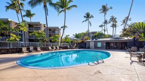 A swimming pool with lounge chairs, palm trees, and a building with a stone facade. - Alii Villas #224 1 Bedroom Condo (Kailua-Kona)