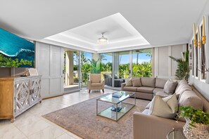 Un salón amplio con una mesa de centro de vidrio, sofá color beige y vista a un jardín tropical a través de grandes puertas correderas de cristal.