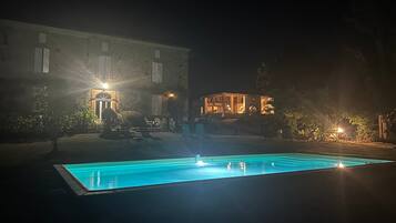 Une vue nocturne d’un bâtiment doté d’une piscine, d’un espace extérieur aménagé et de fenêtres illuminées.