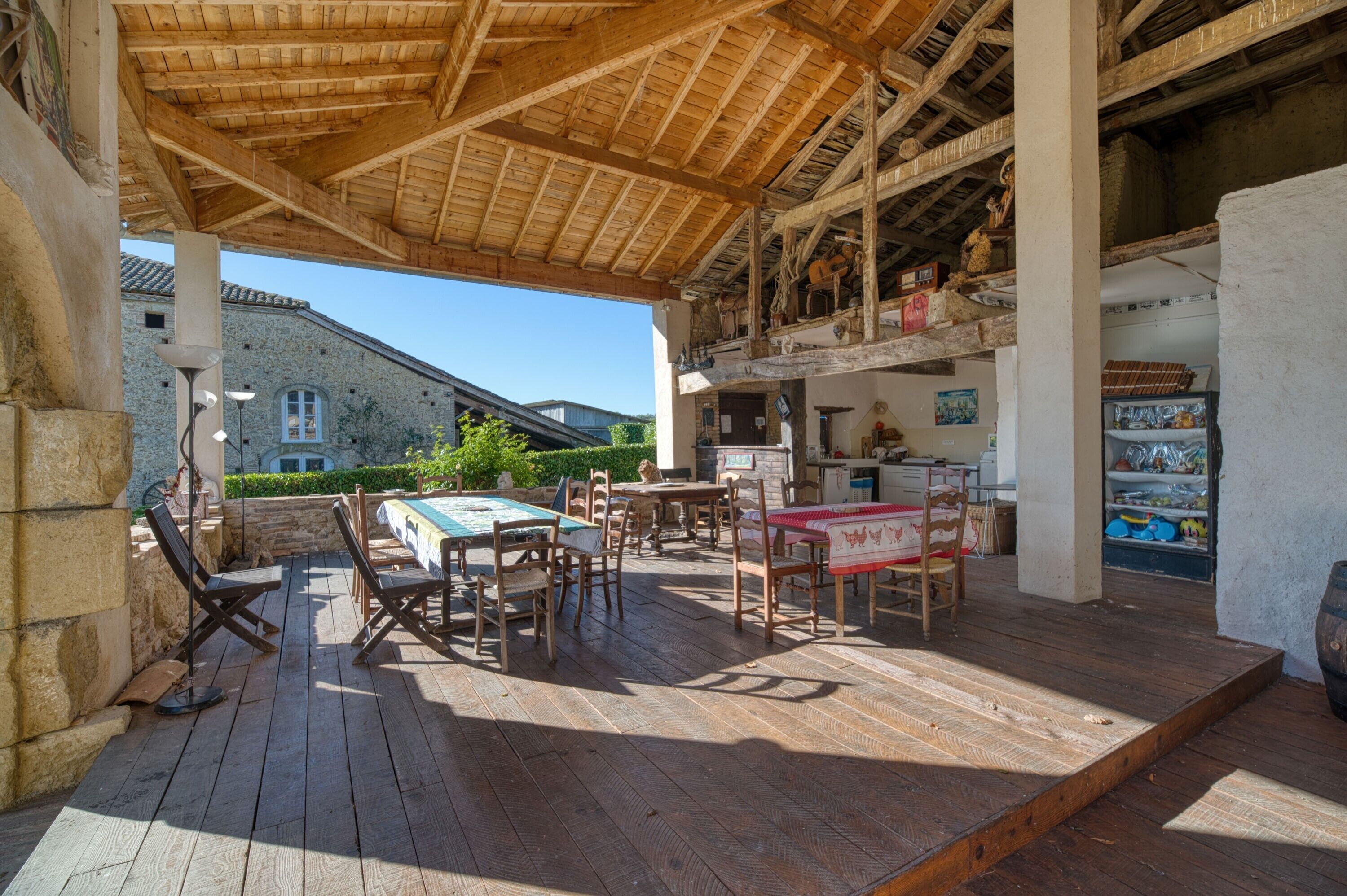 An outdoor wooden deck with tables and chairs, a covered area with a wooden ceiling, and a stone building in the background.