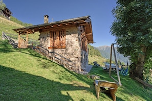 Un bâtiment rustique en pierre, doté d’un escalier en bois menant à une terrasse en bois, le tout entouré d’une végétation luxuriante et avec des montagnes en toile de fond.