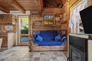 Un intérieur rustique avec des éléments en bois, un canapé bleu, un sol en pierre et une vue sur la verdure à l’extérieur.