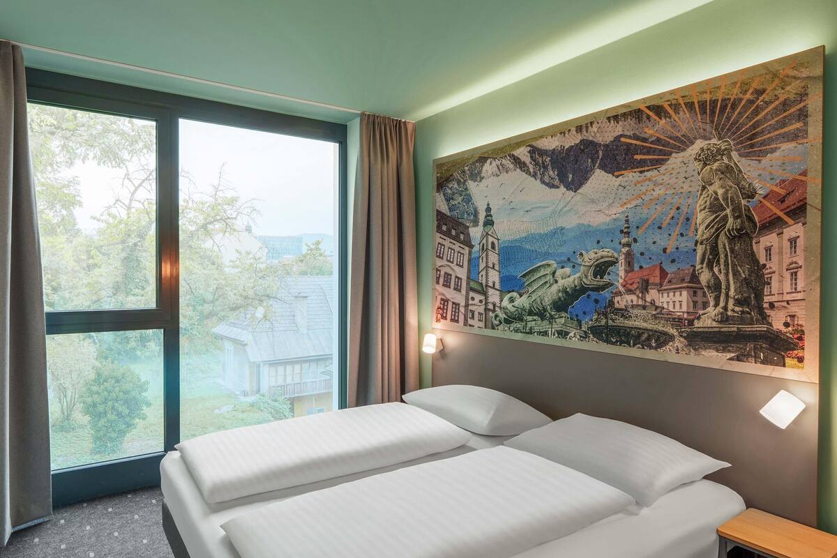 Ein Hotelzimmer mit Bett, Nachttisch, Fenster mit Vorhängen und Wandgemälde.