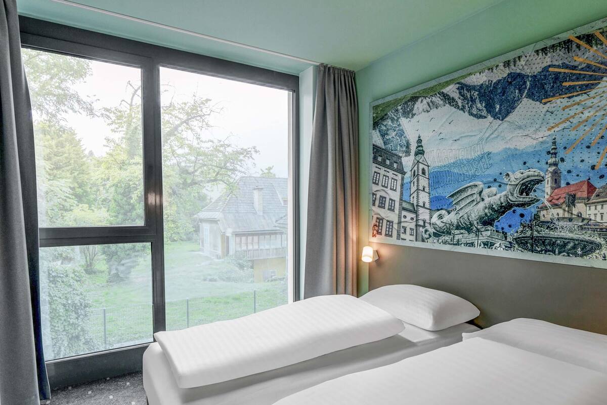 Ein Hotelzimmer mit zwei Betten, einem großen Fenster mit Blick auf ein Haus und Grünflächen, sowie einem Wandgemälde.