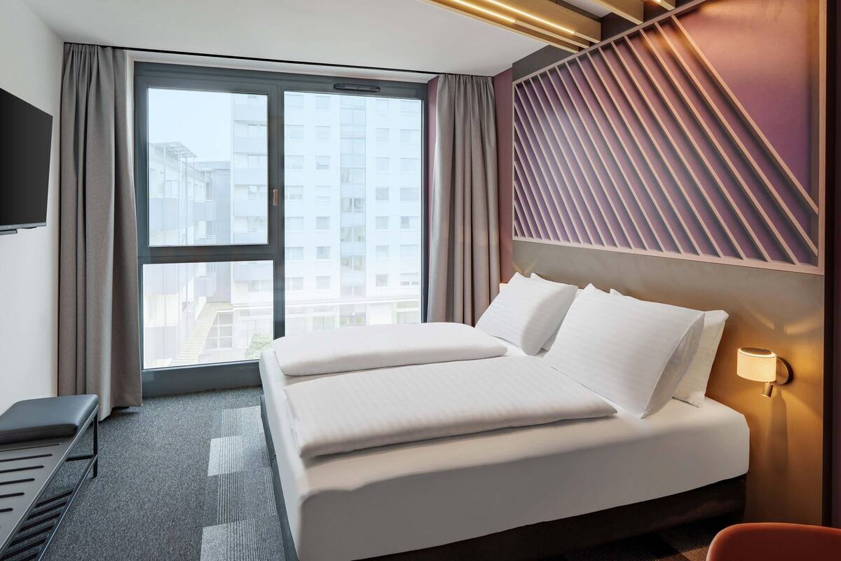 Ein Hotelzimmer mit einem großen Bett, einem Schreibtisch, einem Fernseher und einem Fenster mit Blick auf die Stadt.