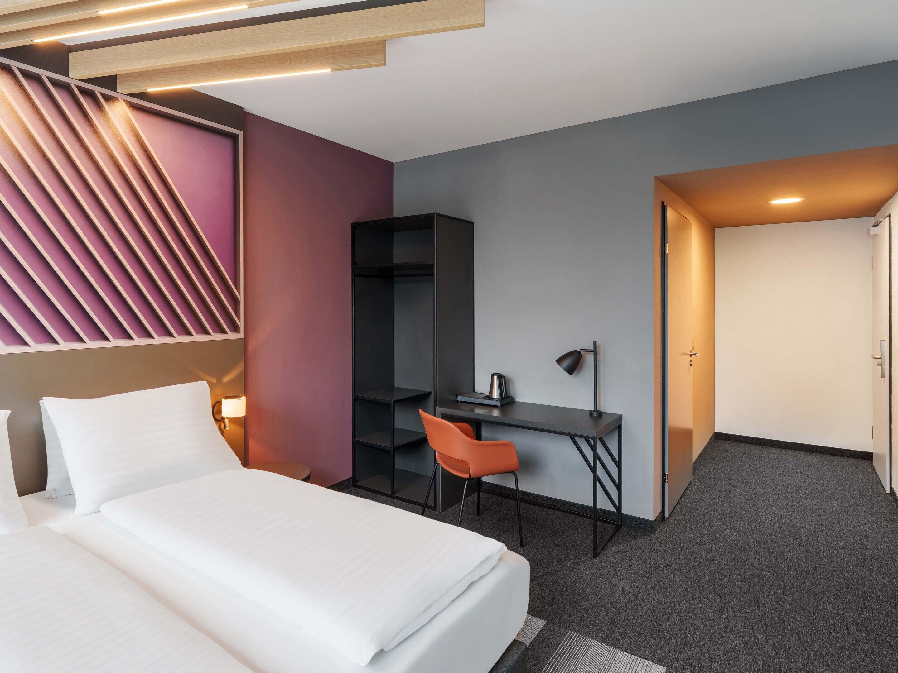 A modern hotel room with a bed, desk, chair, and a wardrobe.