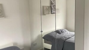 A bedroom with a single bed, a bedside table, and a wall-mounted shelf with decorative items. - Greenwich Oasis Near o2 arena with Spacious Charm (Greenwich)