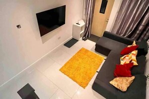 A modern living room with a flat-screen TV mounted on the wall, a dark-colored sofa with red and beige cushions, and a yellow rug. - Greenwich Oasis Near o2 arena with Spacious Charm (Greenwich)