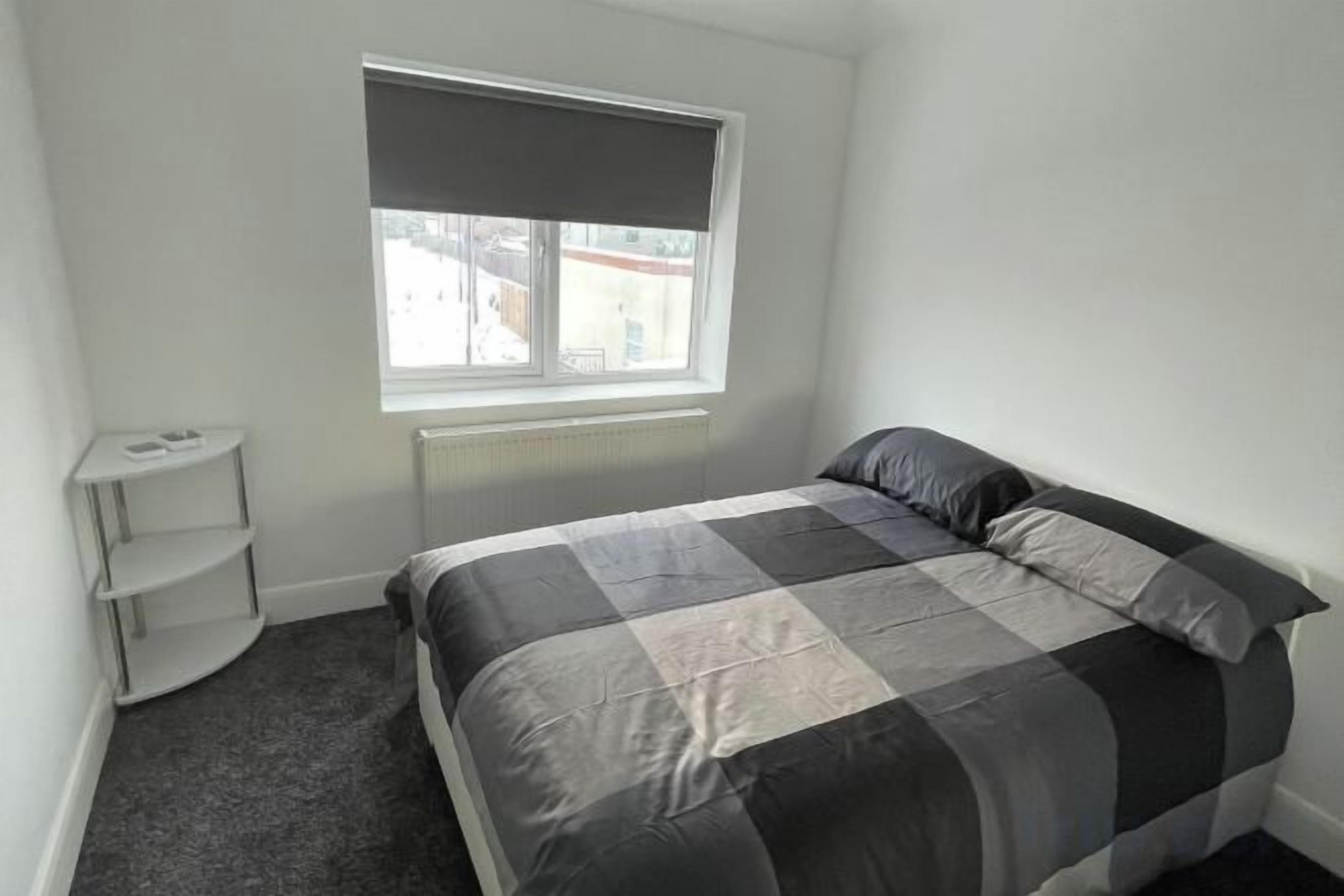 A bedroom with a bed, a window with a blind, a radiator, and a white shelf unit.