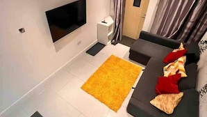 A modern living room with a flat-screen TV mounted on the wall, a grey sofa with red and beige cushions, and a yellow rug on the floor. - Greenwich Oasis Near o2 arena with Spacious Charm (Greenwich)