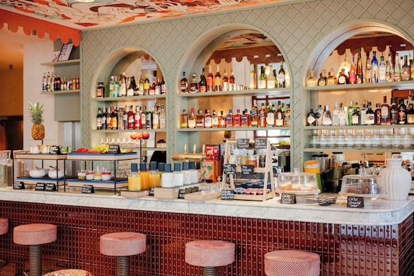 A bar with multiple shelves stocked with various bottles, a fruit display, and a counter with stools. - Hotel Posidonia (Nice)