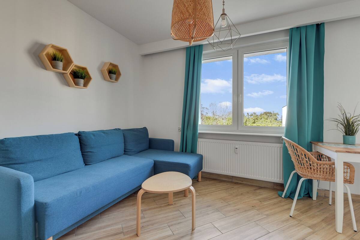 Una sala moderna con un sofá azul, una mesa redonda de madera y una silla de mimbre. Hay tres estantes hexagonales con plantas en la pared, y una ventana con cortinas color turquesa.
